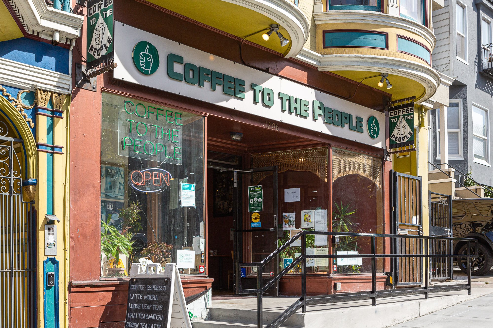 Exterior view of "Coffee to the People" cafe with an open sign and menu board in front, located in a colorful building.