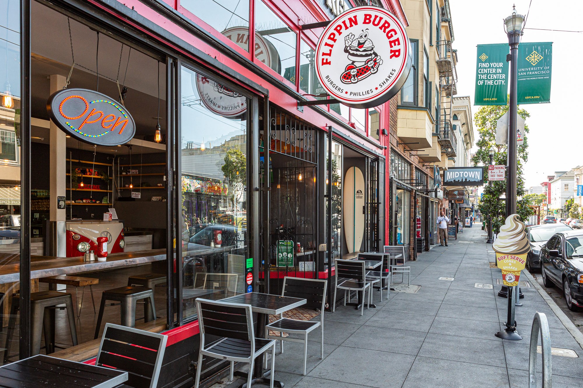Street view of Flippin Burger restaurant with outdoor seating, an "Open" sign, and a menu featuring burgers, Phillies, and shakes. Located on a busy urban sidewalk with other shops and street parking.