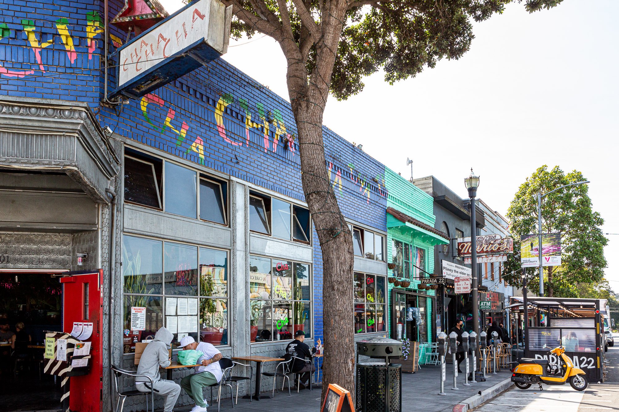 Street view of a bustling urban scene with colorful buildings and outdoor seating. People are sitting at tables outside a blue building with vibrant graffiti. A scooter is parked in front, and there's a tree and lamppost lining the street.