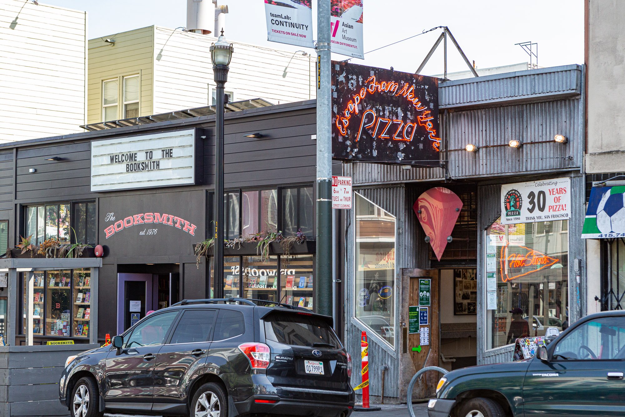 Street view of The Booksmith bookstore and Escape from New York Pizza with neon signs and parked cars in front on a sunny day.