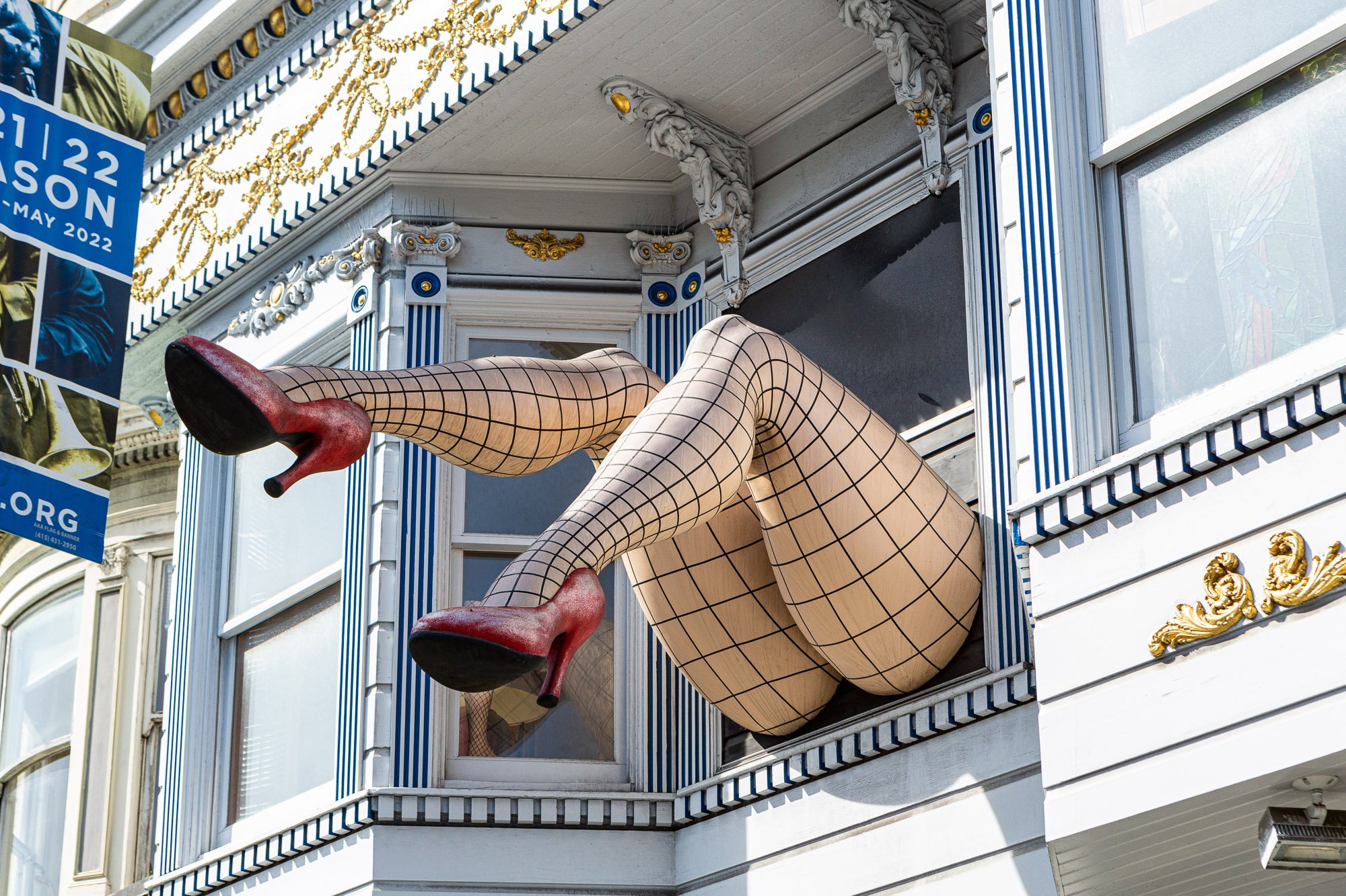 Decorative mannequin legs wearing fishnet stockings and red high heels protruding from a window of a building with ornate trim.