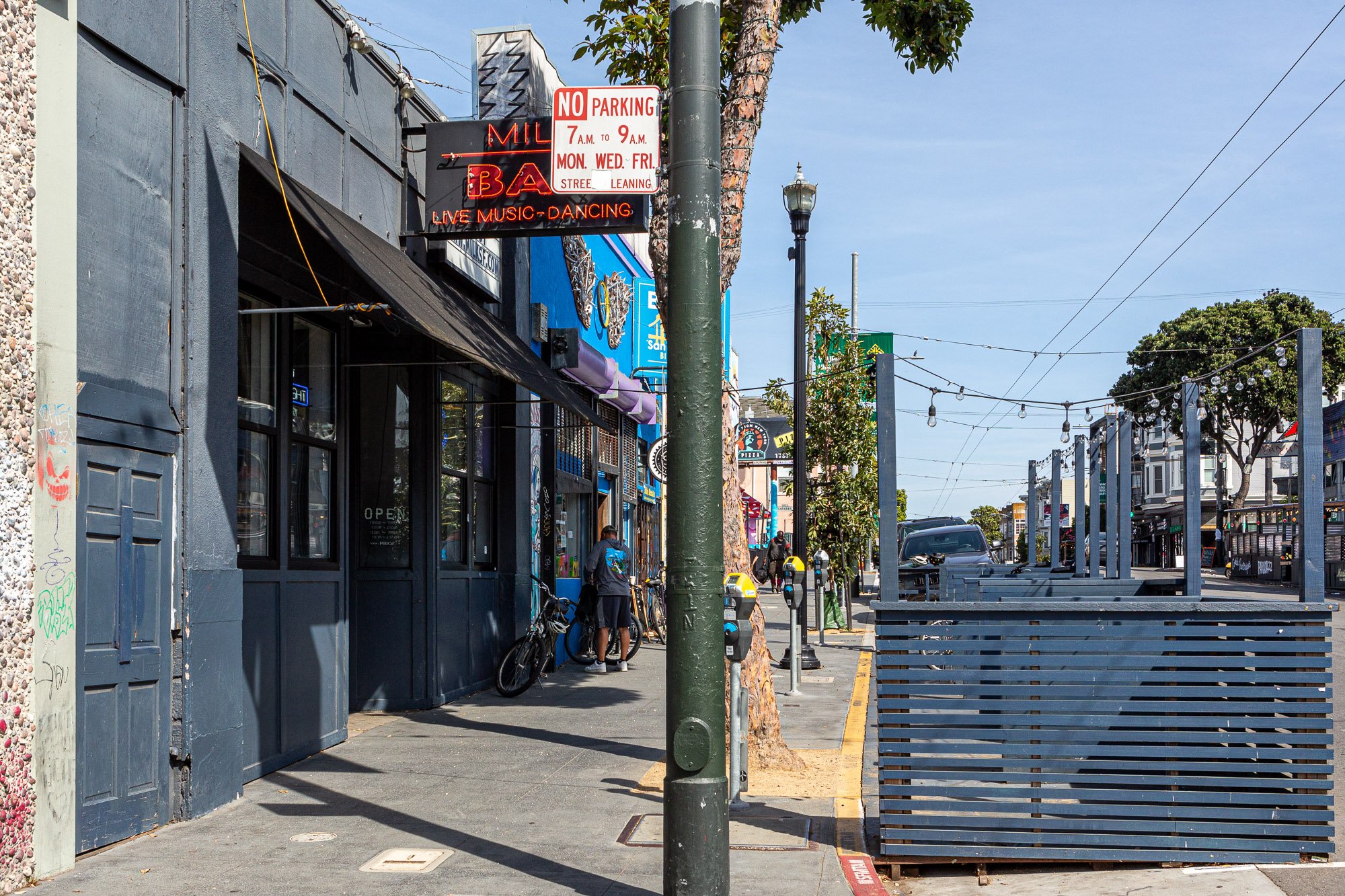 Street scene in an urban area with a bar sign for live music, bicycles parked, and outdoor seating area. Visible no parking sign, people walking, and trees along the sidewalk. Overhead string lights are hung across the street.