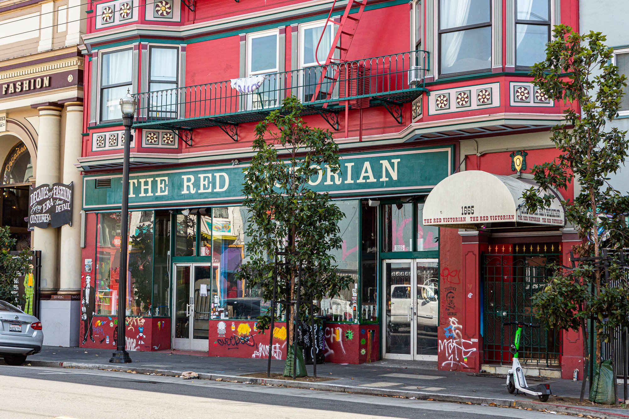 Street view of "The Red Victorian," a historic red and green building with balconies, decorative details, and graffiti at the entrance, located next to a store sign reading "Decades of Fashion."