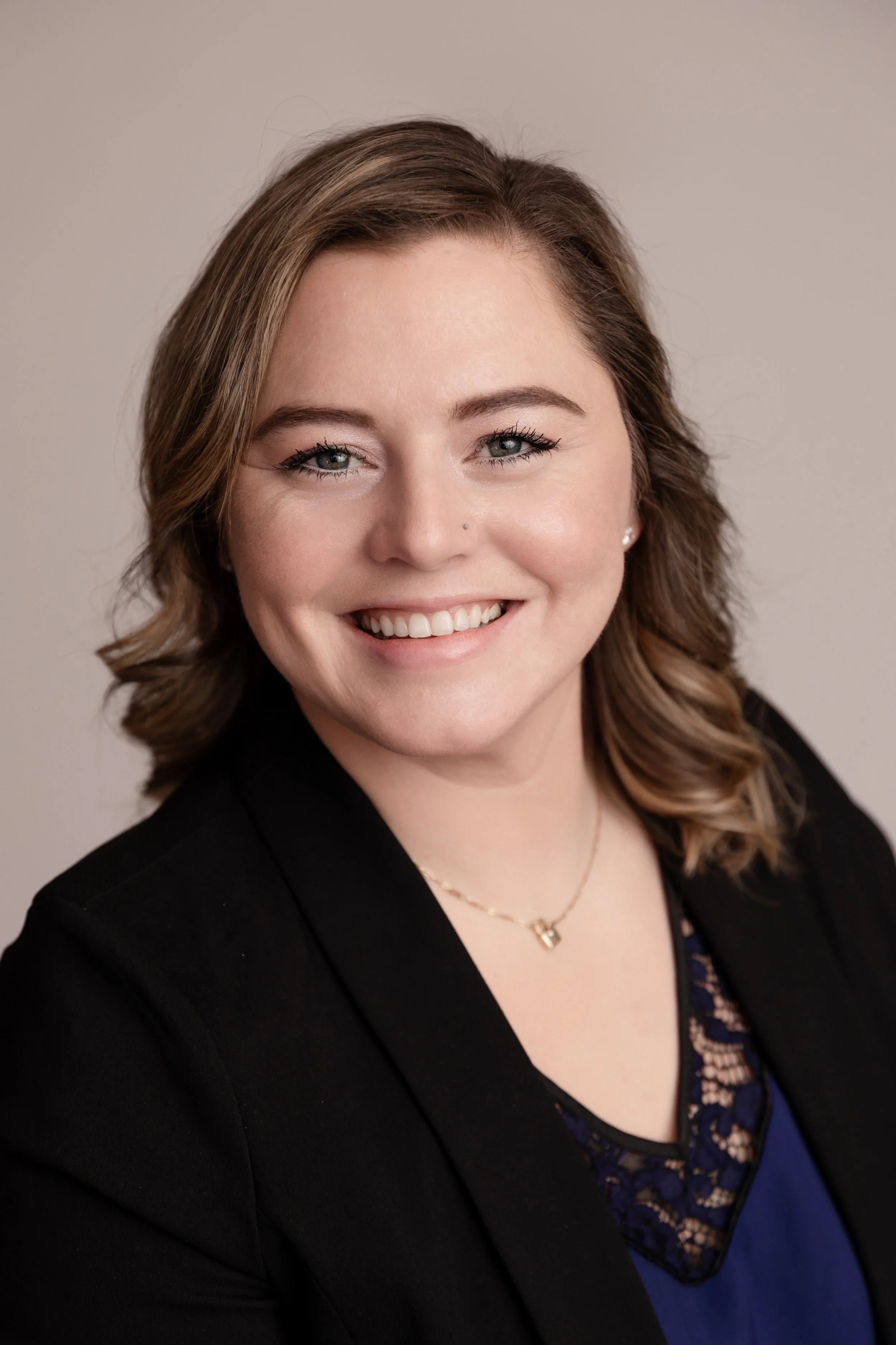 Portrait of a woman with light skin, wavy brown hair, blue eyes, and a smile, wearing a black blazer and a blue top with lace detail, against a neutral background.