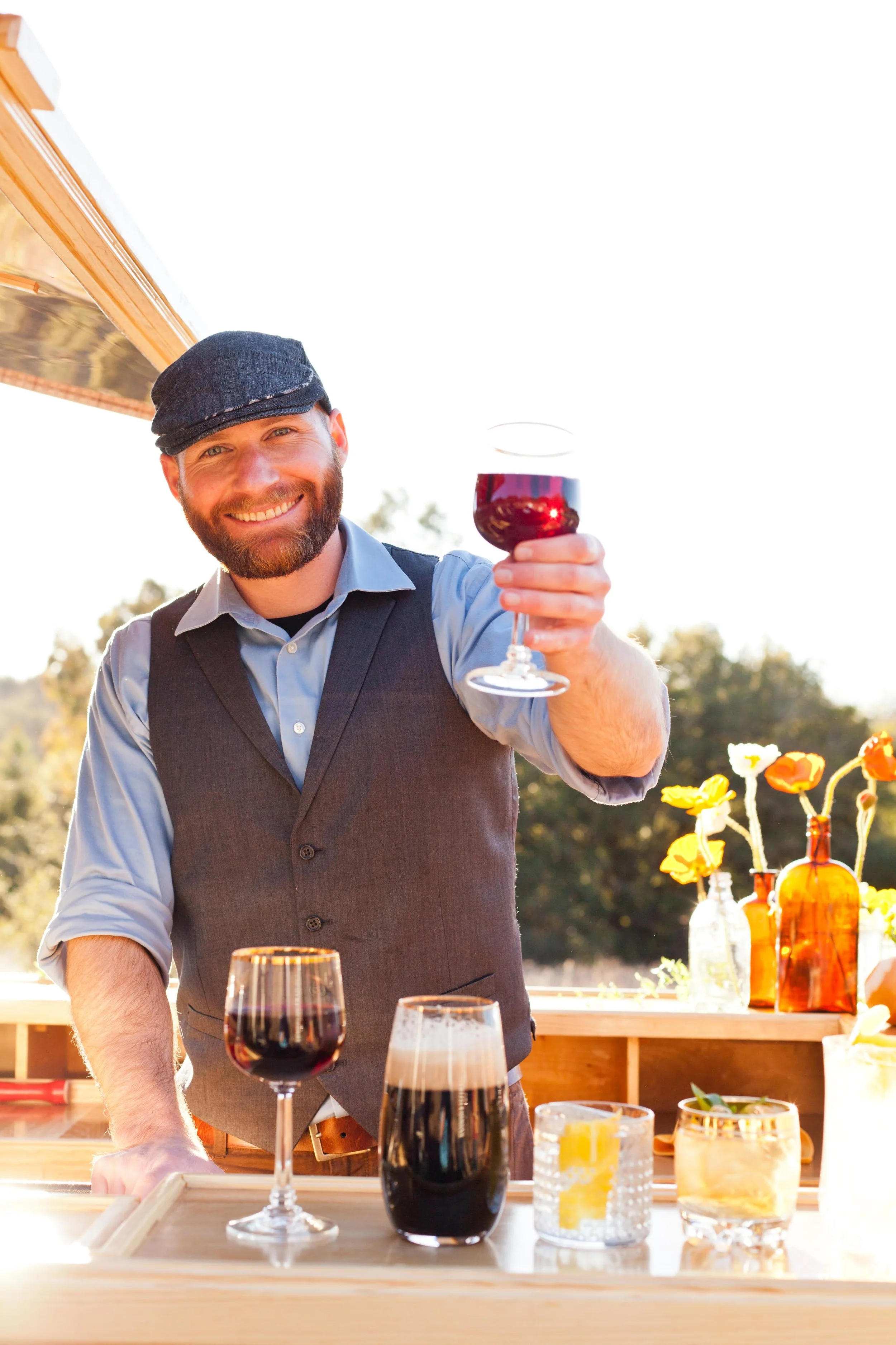 Smiling man in a vest and cap raising a glass of red wine outdoors, with various drinks on a table in front, against a sunny background with trees and flowers.