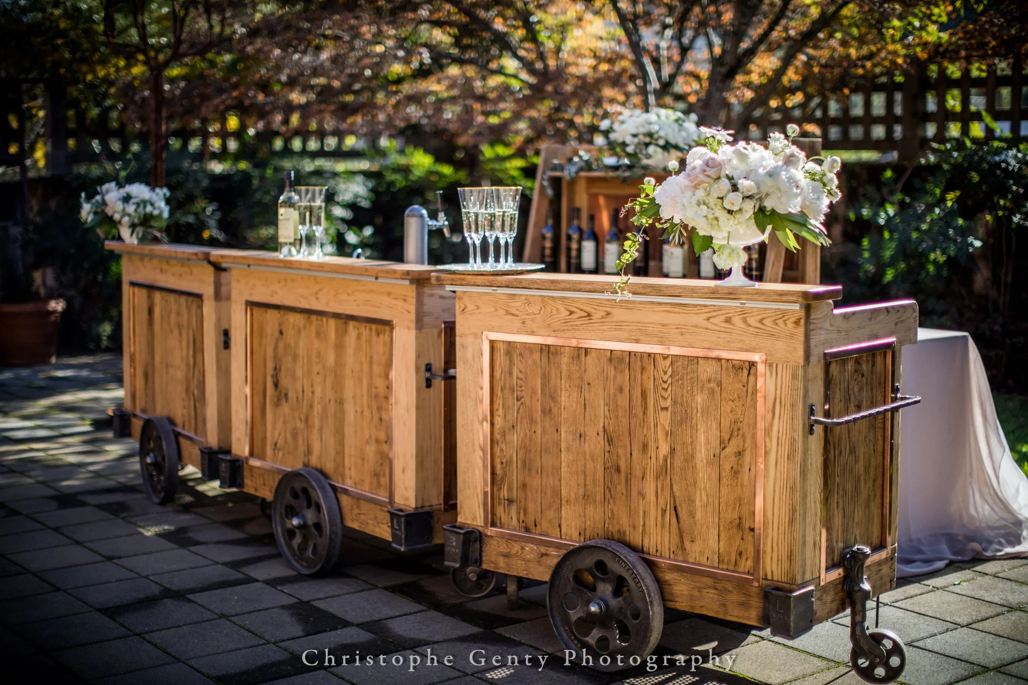 A wooden outdoor bar cart with wheels, decorated with white flowers and glassware, set in a garden with trees and bushes.