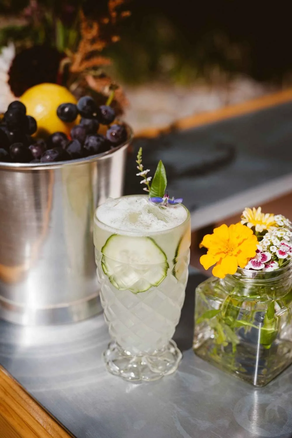 A glass of cucumber-infused cocktail garnished with lavender and greenery, next to a vase with yellow and pink flowers, and a metal bucket filled with blueberries and a lemon in the background.