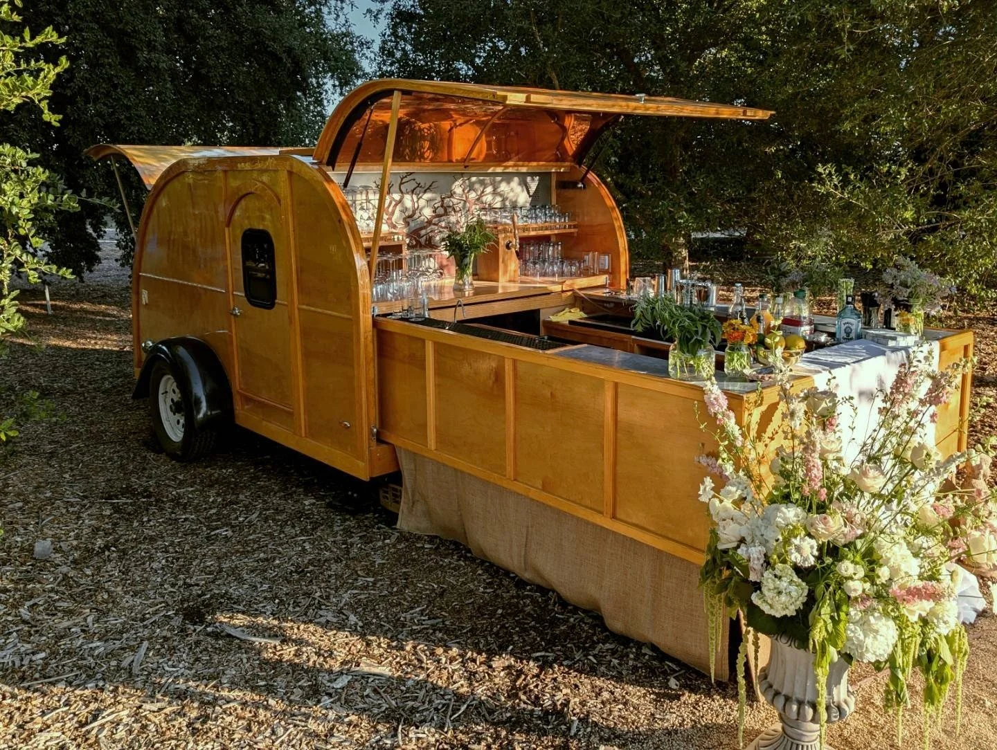 A wooden mobile bar trailer with an open top, set outdoors among trees, decorated with flowers and glassware, with a floral arrangement in a tall white vase in front.