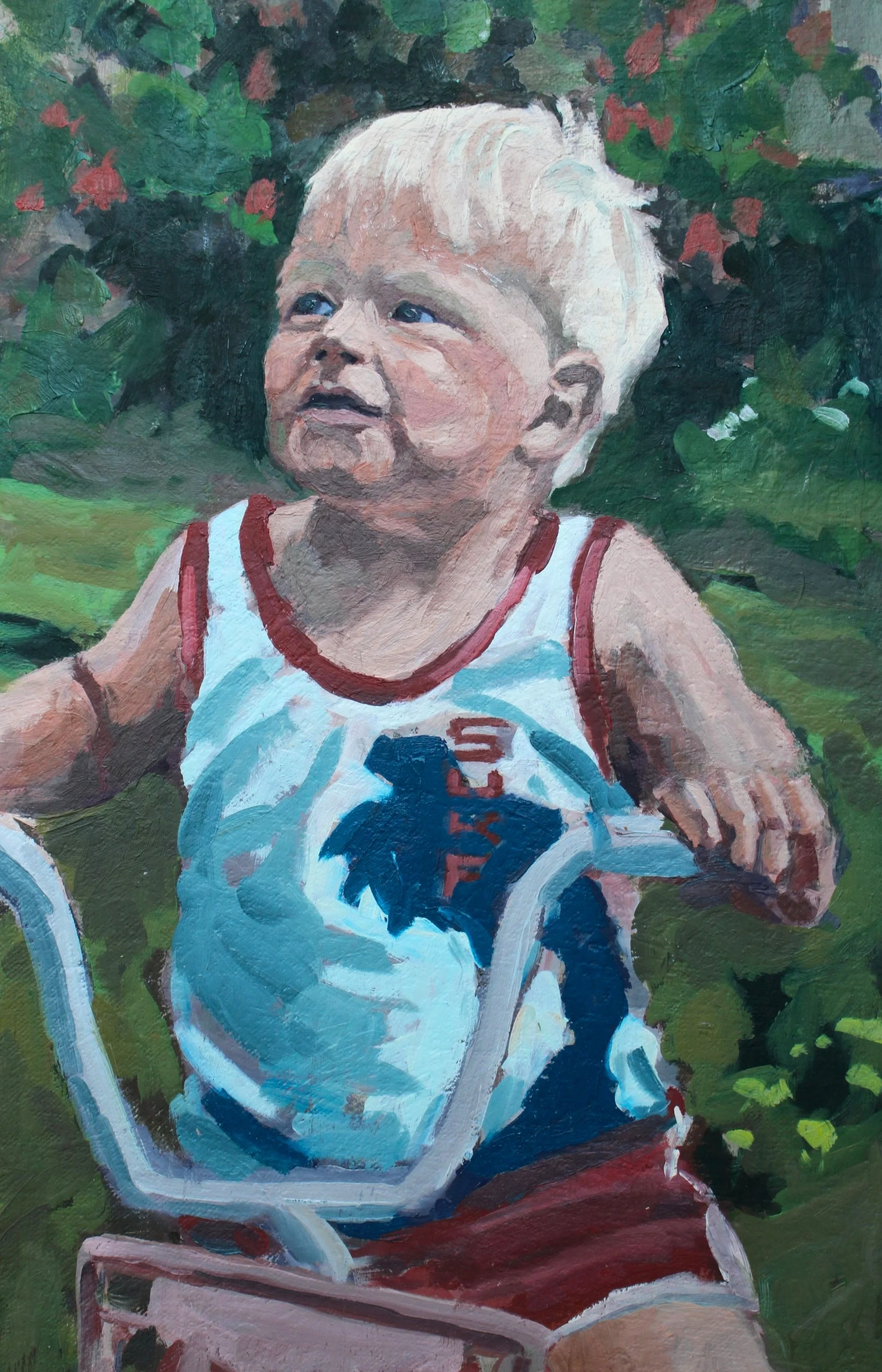 A young child with blonde hair and blue eyes sitting outside, wearing a sleeveless shirt with a large bat symbol on it, surrounded by green foliage.