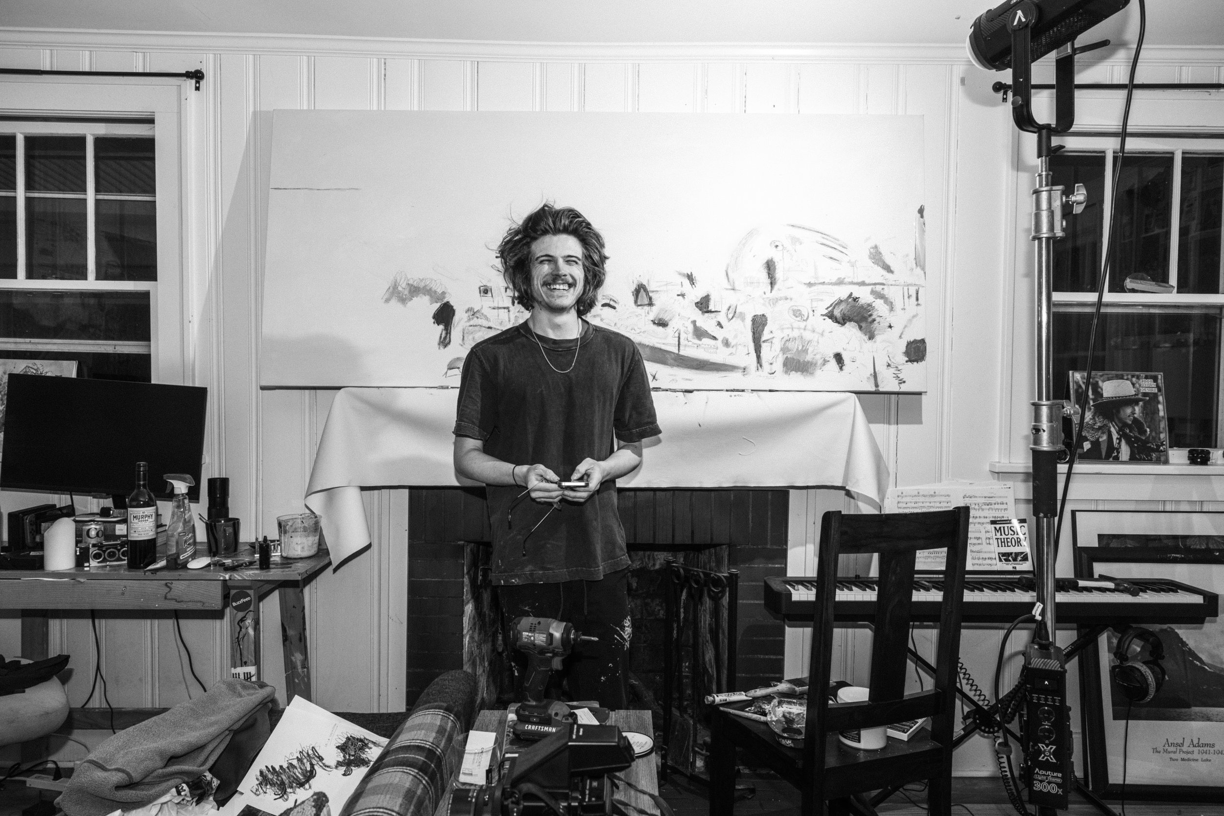 A young man with curly hair and a mustache smiling, standing in a cluttered room with art supplies, a television, and a keyboard, in front of a large abstract painting.