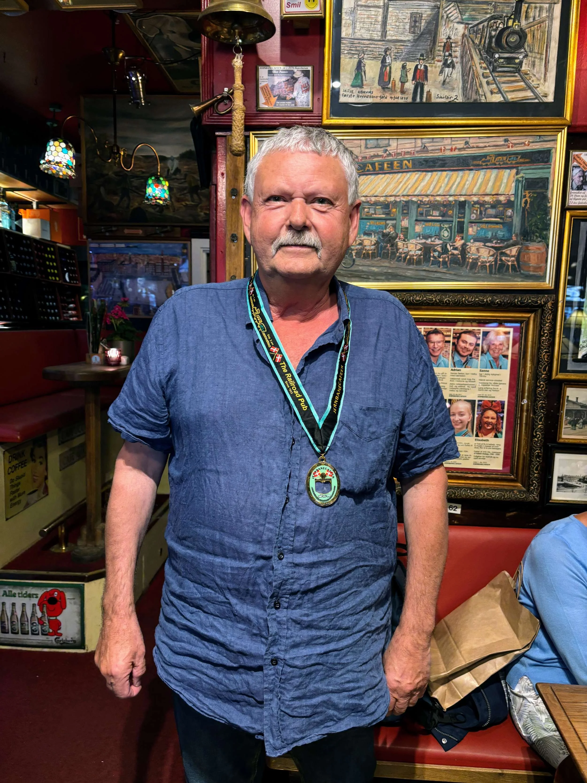 En ældre mand med hvidt hår og skæg står i en restaurant, bærer en medalje om halsen, omringet af billeder og kunstværker på væggen.