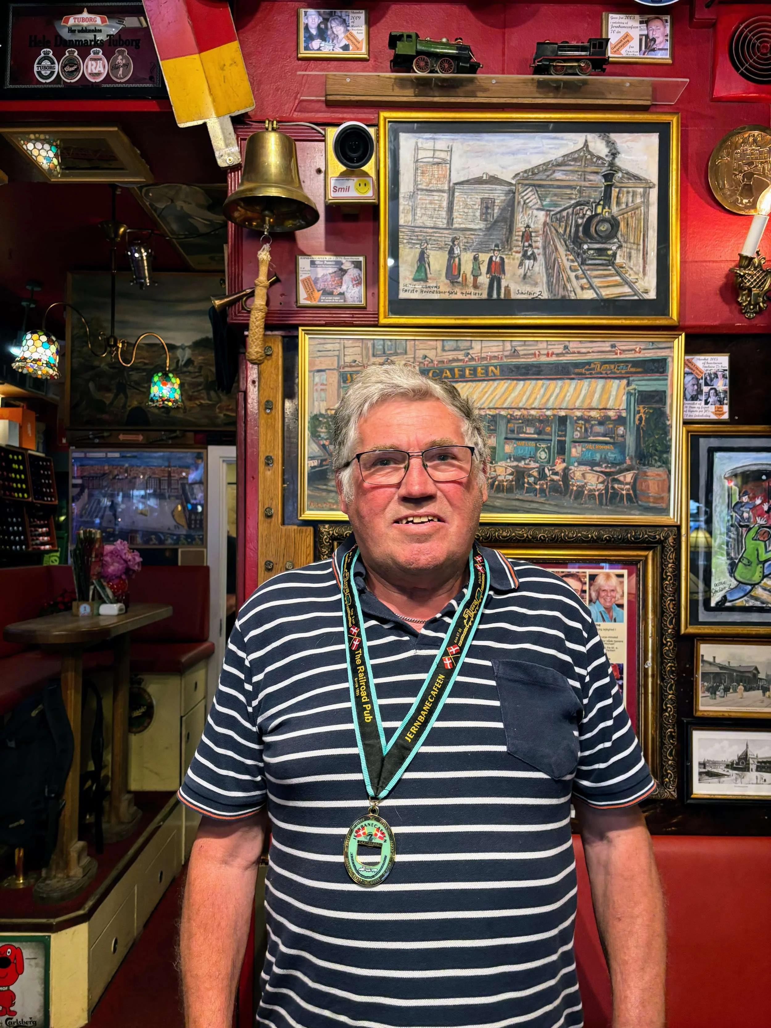 En mand står foran en væg med malerier og memorabilia i en restaurant eller pub, iført en stribet polo og en medalje om halsen.