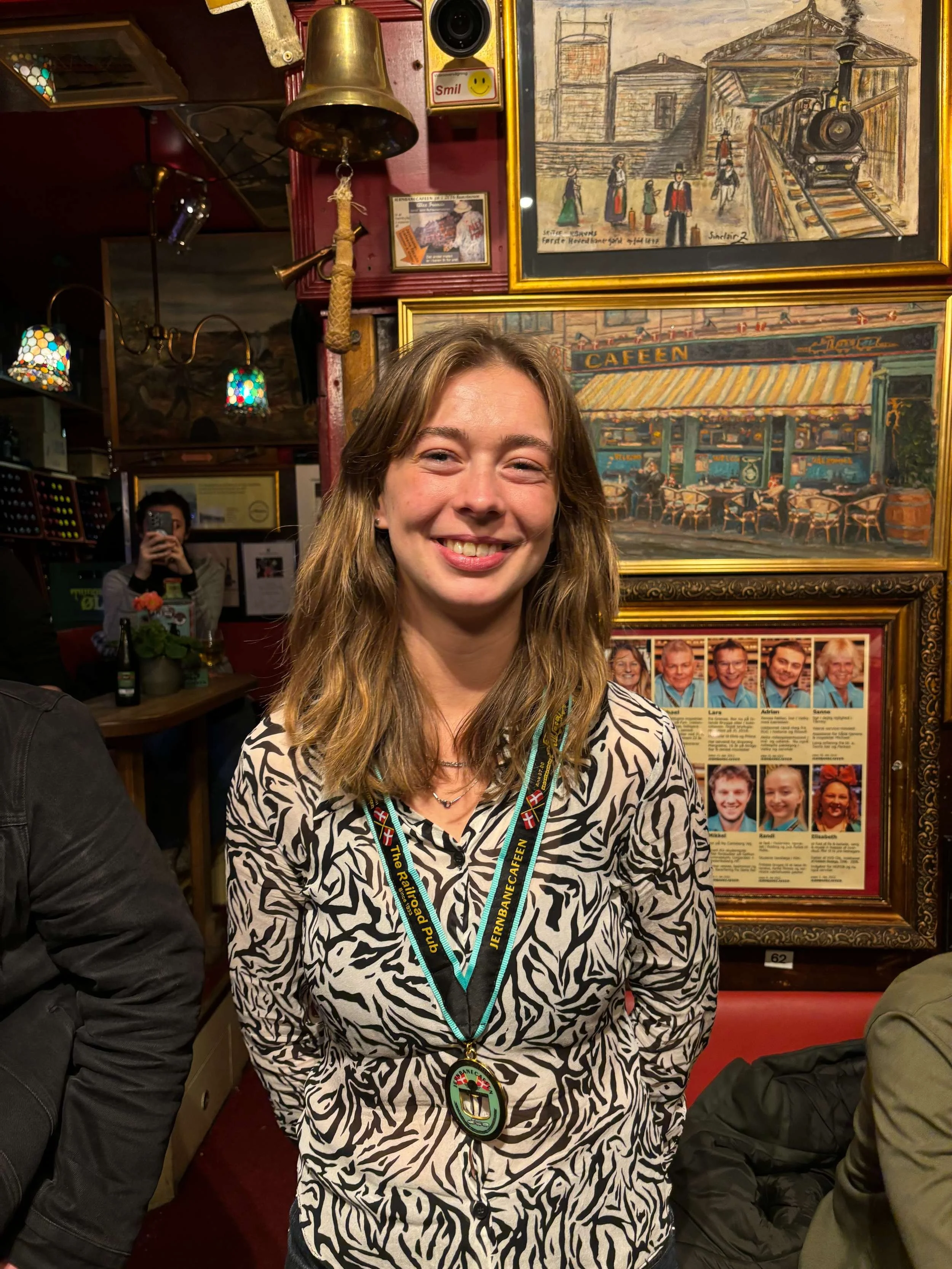En smilende kvinde med brunt, bølget hår, iført en zebra-printet skjorte og en medalje rundt halsen, sidder i en hyggelig cafe med kunst på væggene.