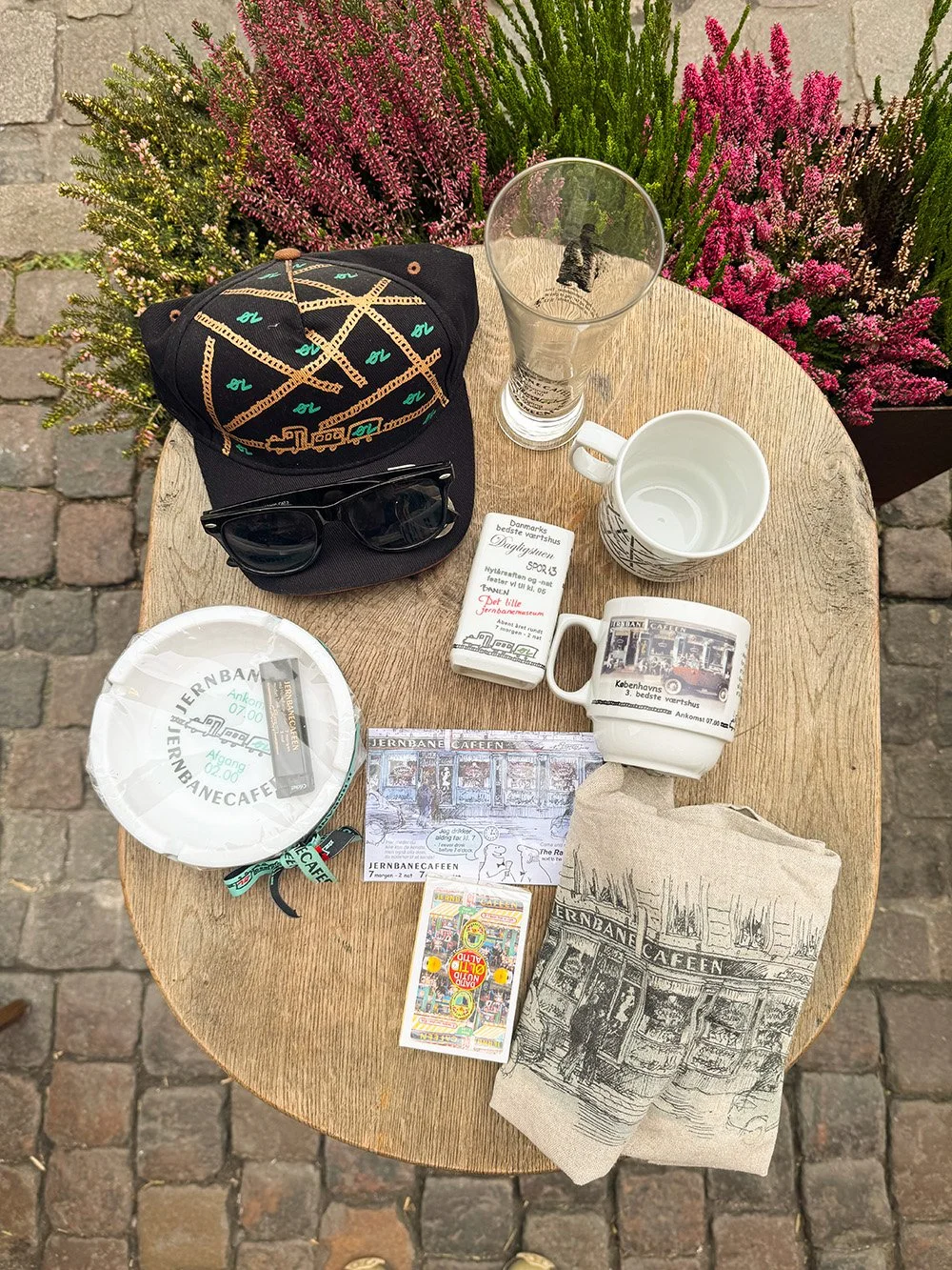 Bord med diverse souvenirs og personlige genstande foran blomster og brosten.