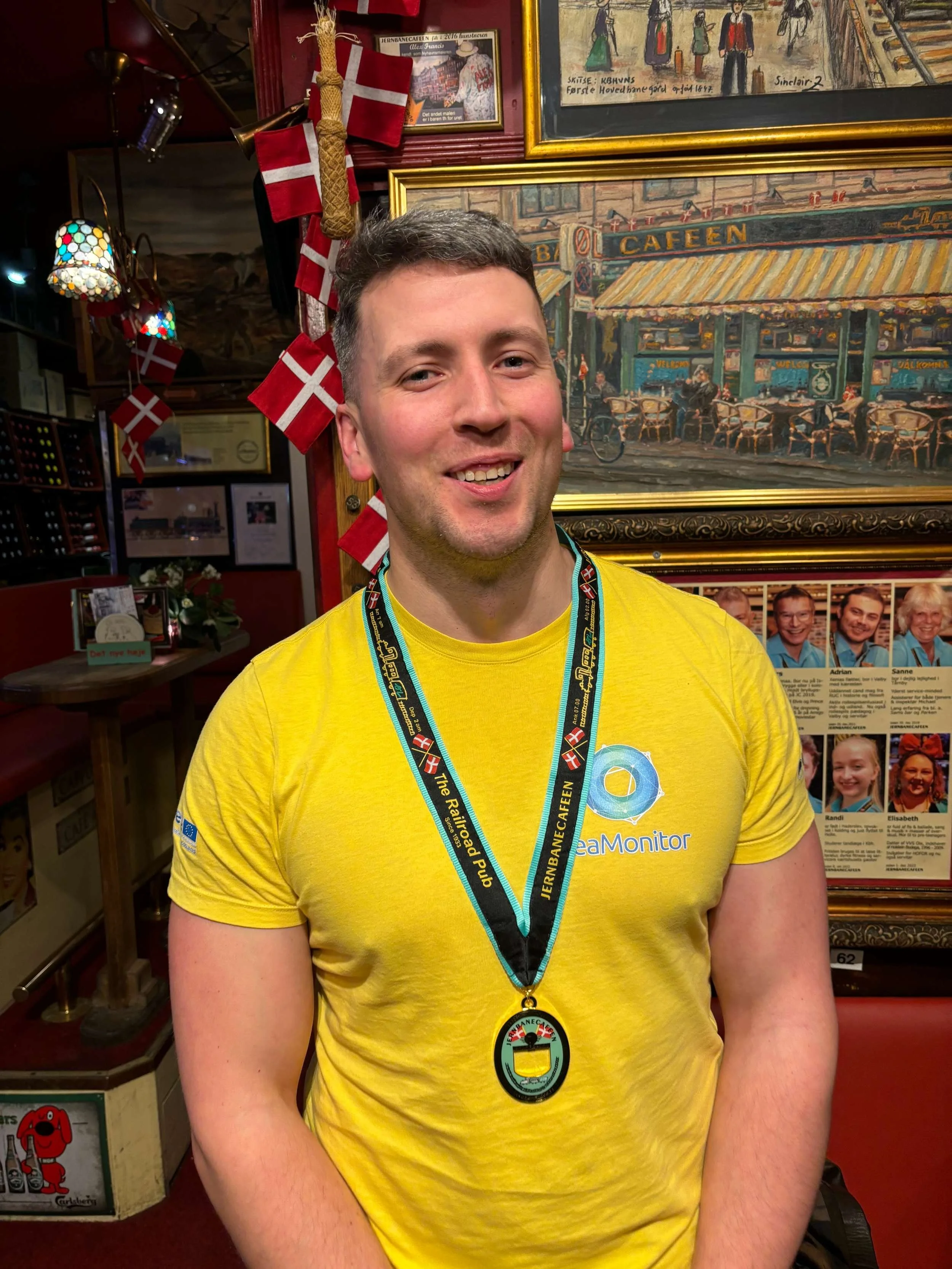 En smilende mand med kort mørkt hår iført en gul T-shirt med et logo og en medalje rundt halsen, stående indendørs blandt vægdekorationer og danske flag.