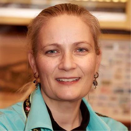 En kvinde med blond hår og blå øjne, smilende, iført en lyseblå uniform og øreringe, i et indendørs miljø.