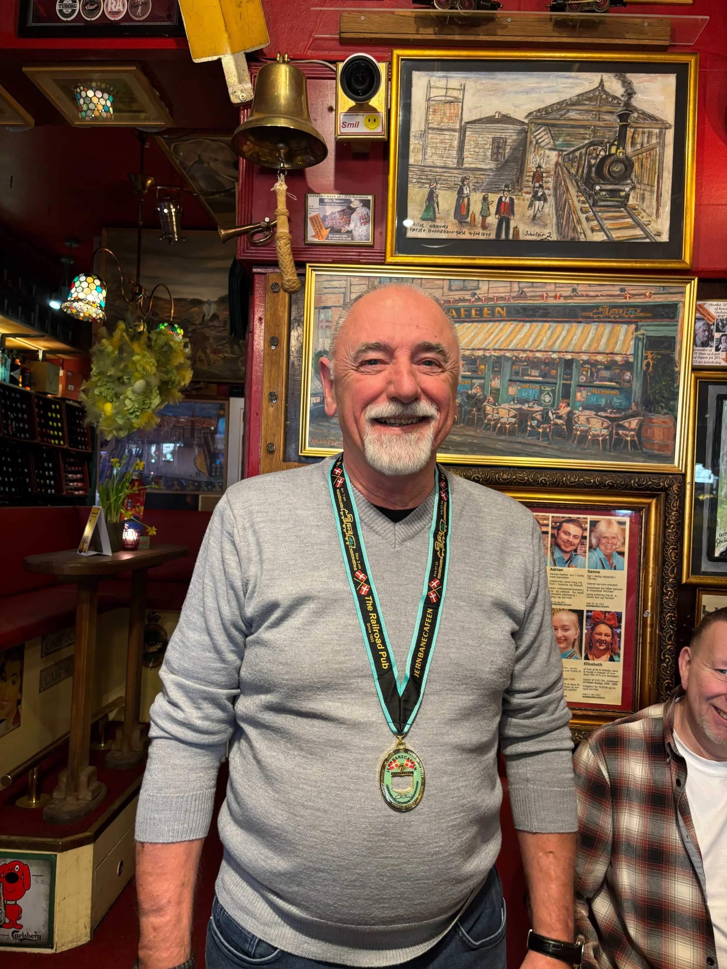 En glad ældre mand med gråt skæg og brunt hår iført en lys grå sweater og medalje rundt om halsen står i en hyggelig indendørs atmosfære med vægdekorationer og malerier. Der er en anden person delvist synlig til højre, og bag ham er der billeder og p
