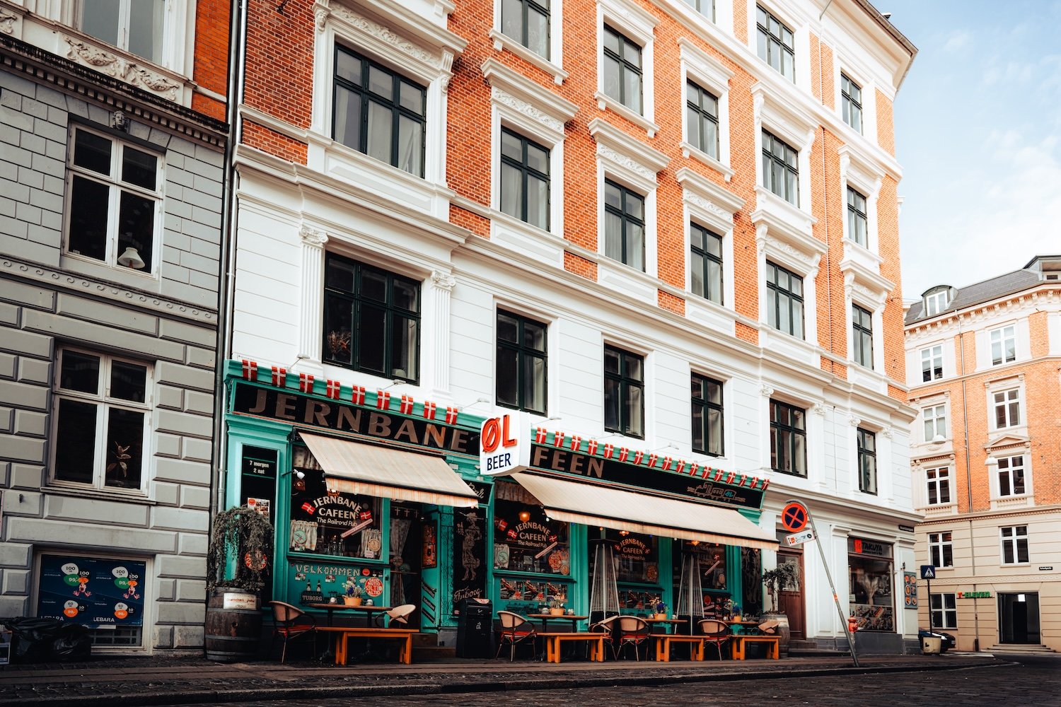 En farvet pub med udendørs siddepladser, der har skilt med teksten 'Jernbane Cafeen', og et mindre skilt med 'øl' og 'øl'.