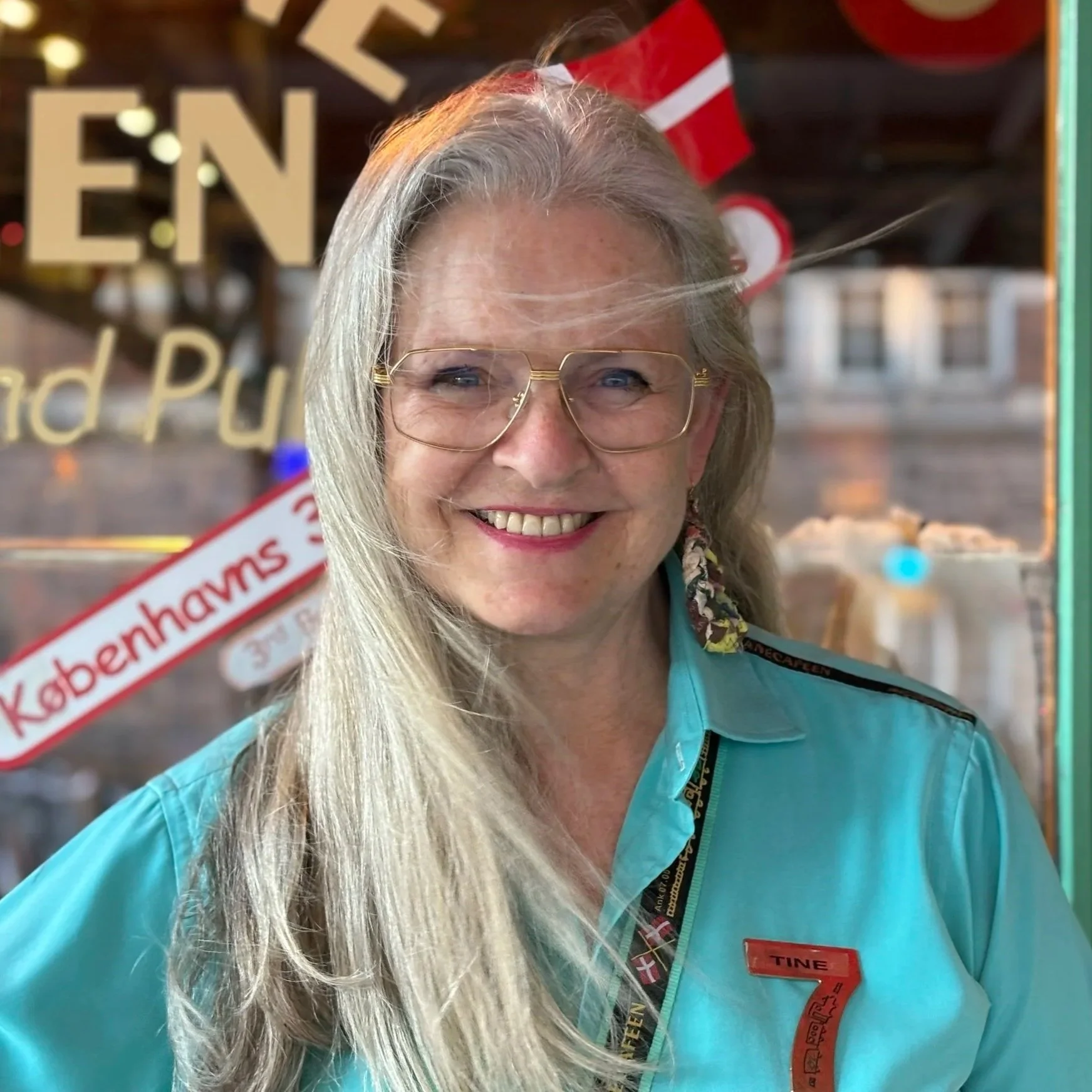 En kvinde med langt blond hår og store brilleglas, iført en lys blå skjorte med navnede Tine, smiler foran en glasdør.