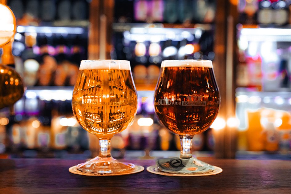 To glas øl på en barbord med bagkamera, med en baggrund af hylder med forskellige varer og lys.