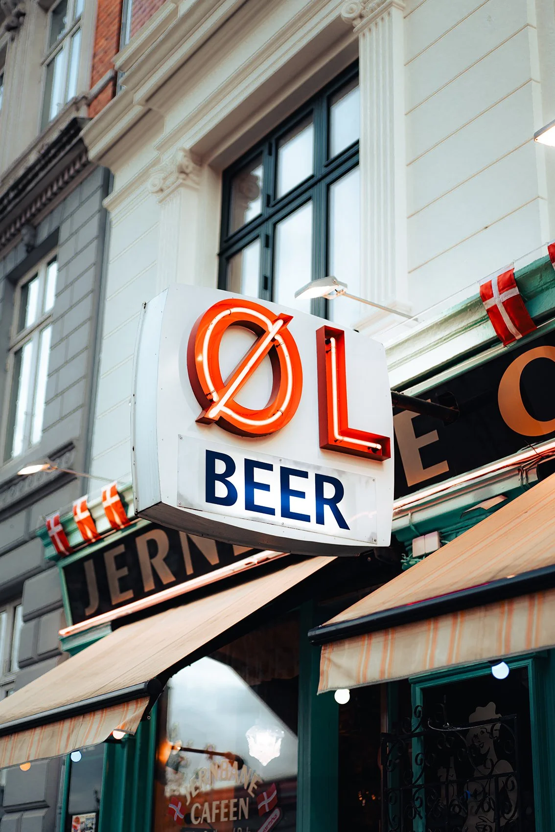Neonskilt med teksten "ØL BEER" på en bygning udendørs, med et café-udendørsområde og en beige stofmarkise.