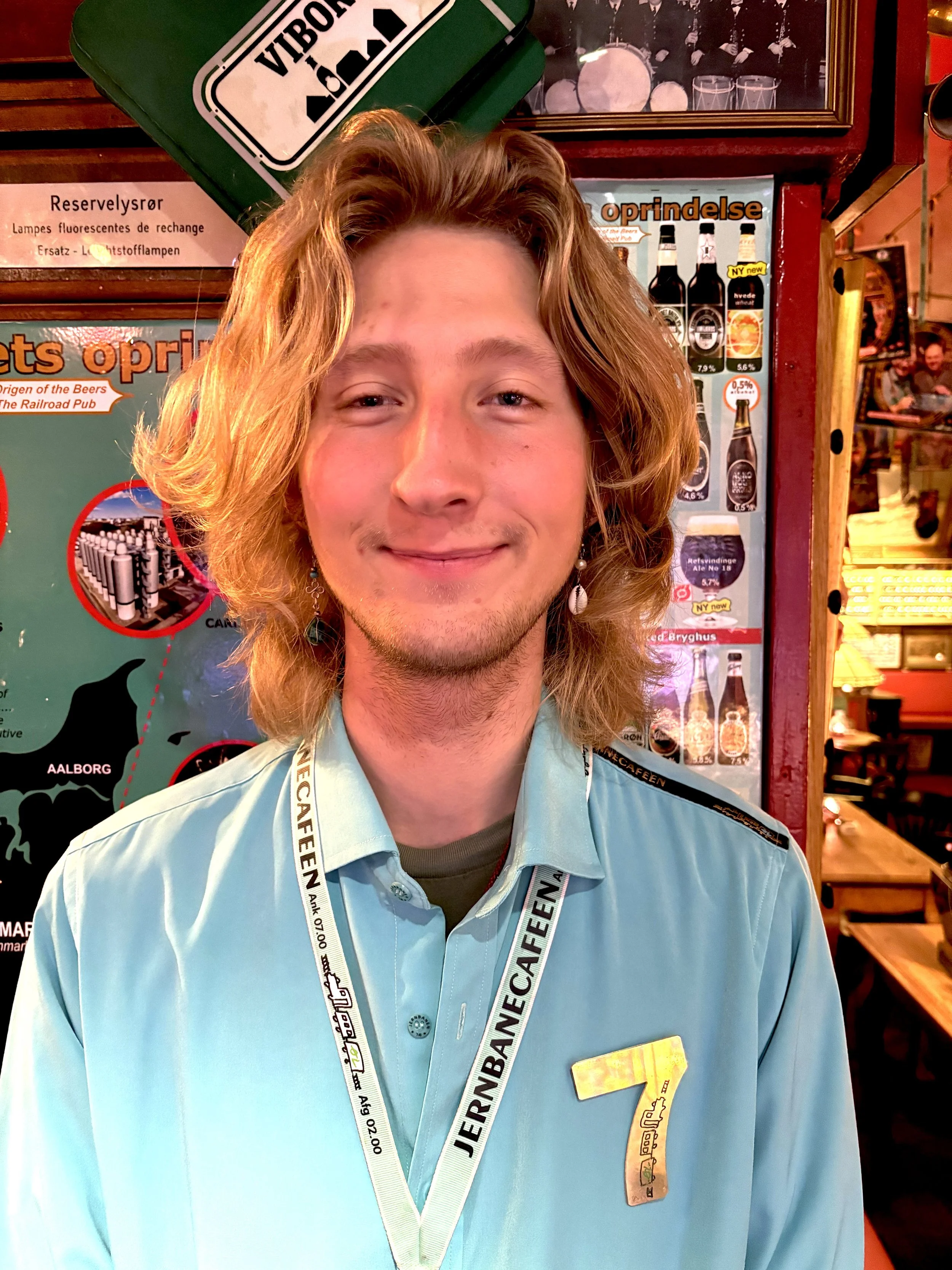 Ung mand med lang, blond hår, iført en lys blå uniform med en gulnummer 7 og et navnemblem, står inde i en restaurant eller bar.