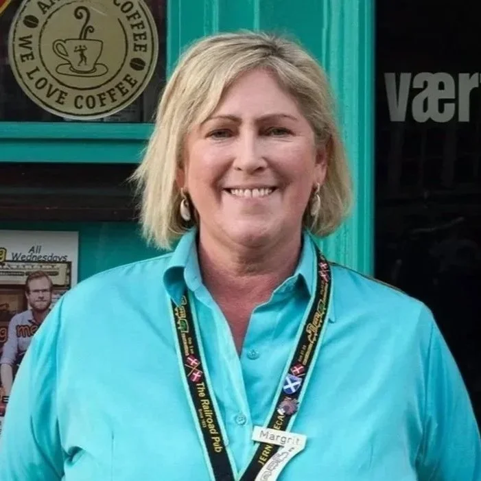 En smilende kvinde med blond hår iført en turkis skjorte og en badge. Baggrund med en café-menu og en grøn dør.