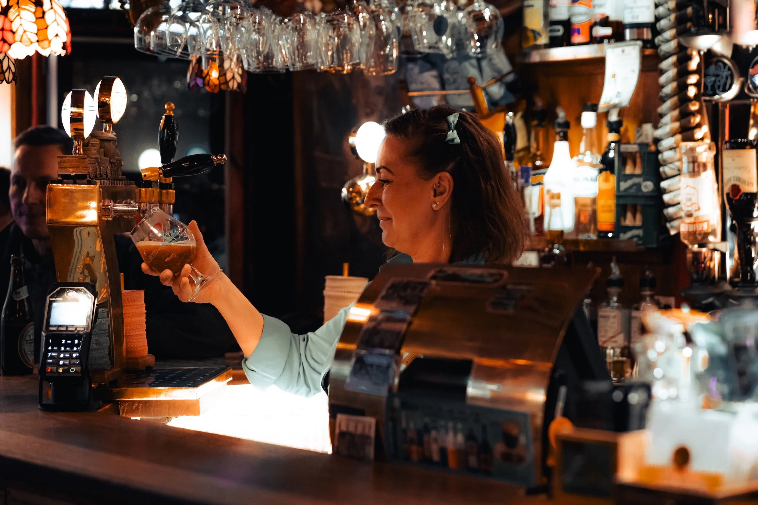 En kvinde smiler, mens hun serverer et glas øl på en bar, omgivet af flasker og barudstyr i en hyggelig, mørk bar.