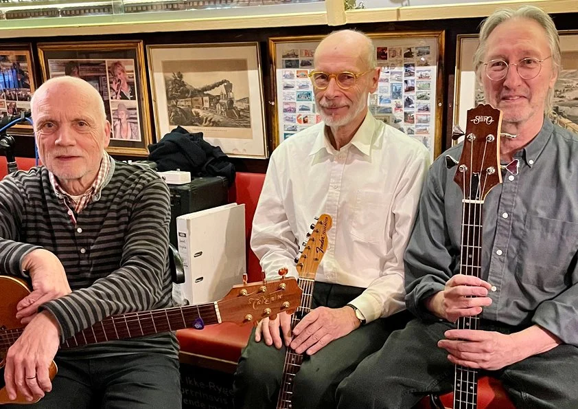 Tre ældre mænd sidder med guitarer i en privat i butik eller bar, omgivet af billeder og plakater på væggen.