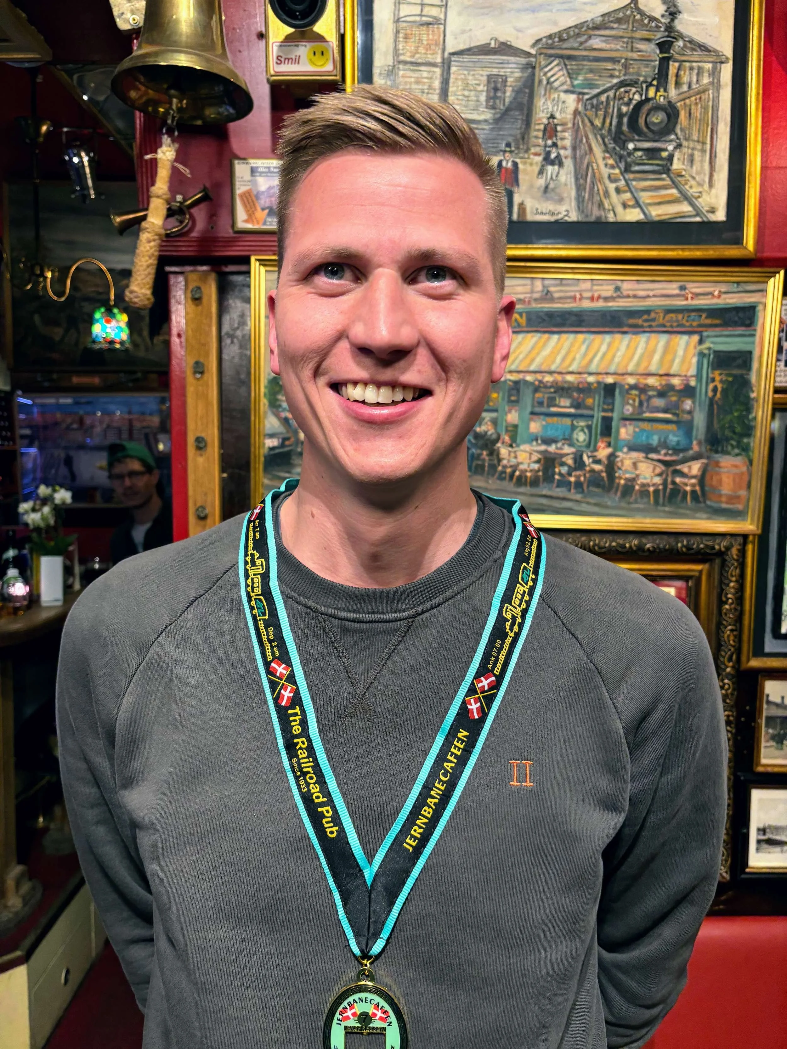En mand med kort, lyst hår smiler foran en væg med billeder og kunstværker, han har en medalje omkring halsen og et medaljebånd med teksten 'JERNBANEFÆN' og 'The Railroader Pub'.