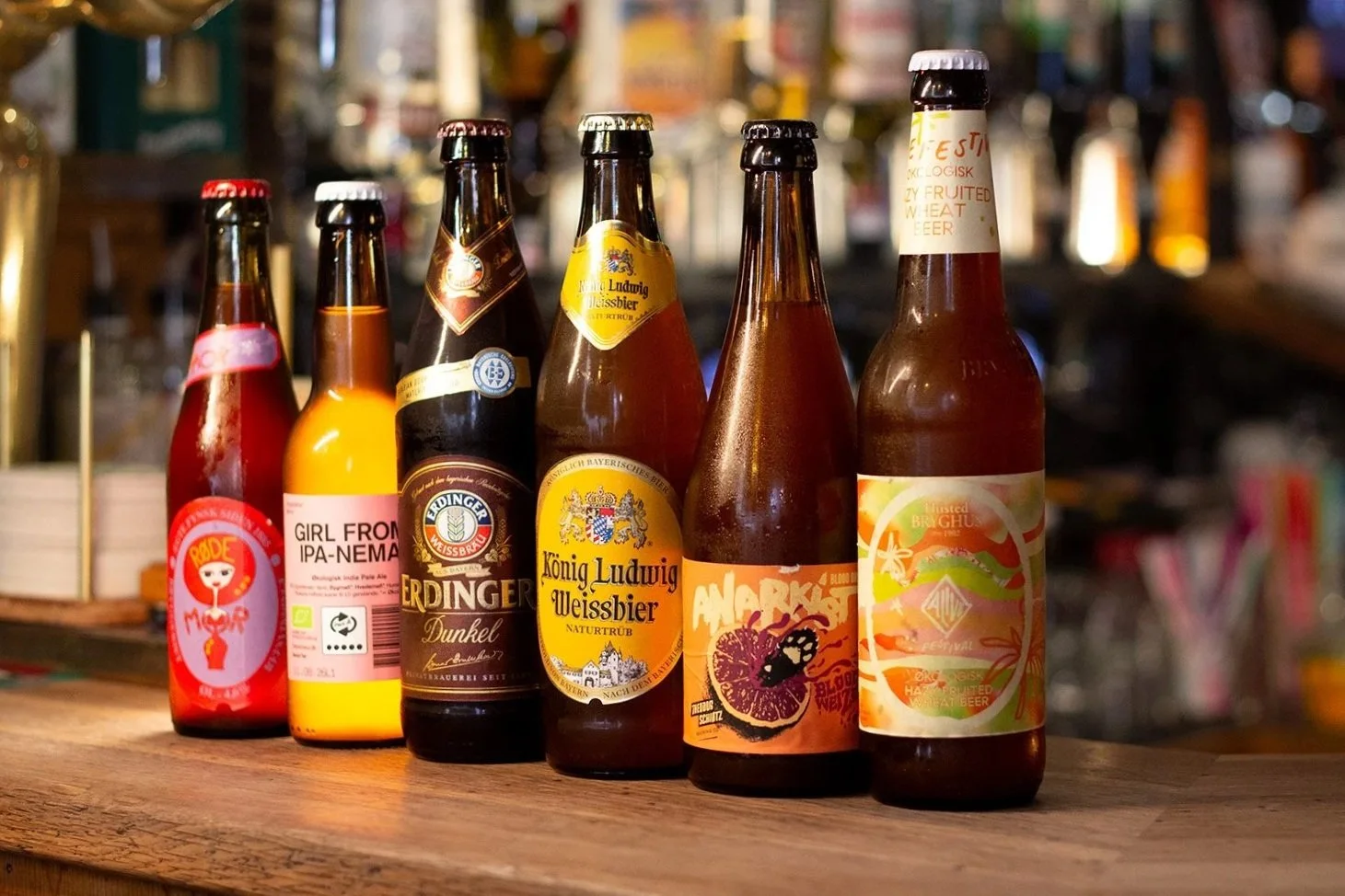 Billedet viser seks forskellige flasker øl på en træbord med en baggrund af en bar.