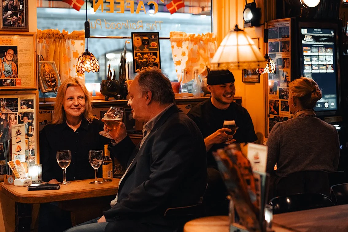 Tre personer sidder i en hyggelig restaurant med varme belysning og dekorationer, og nyder drinks. En kvinde og to mænd, hvoraf den ene holder et glas øl.