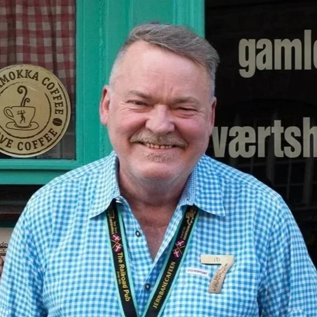 En glad mand med uniform og en badge, der står foran en grøn dør og en vindue med tekst og logo for en kaffebutik.