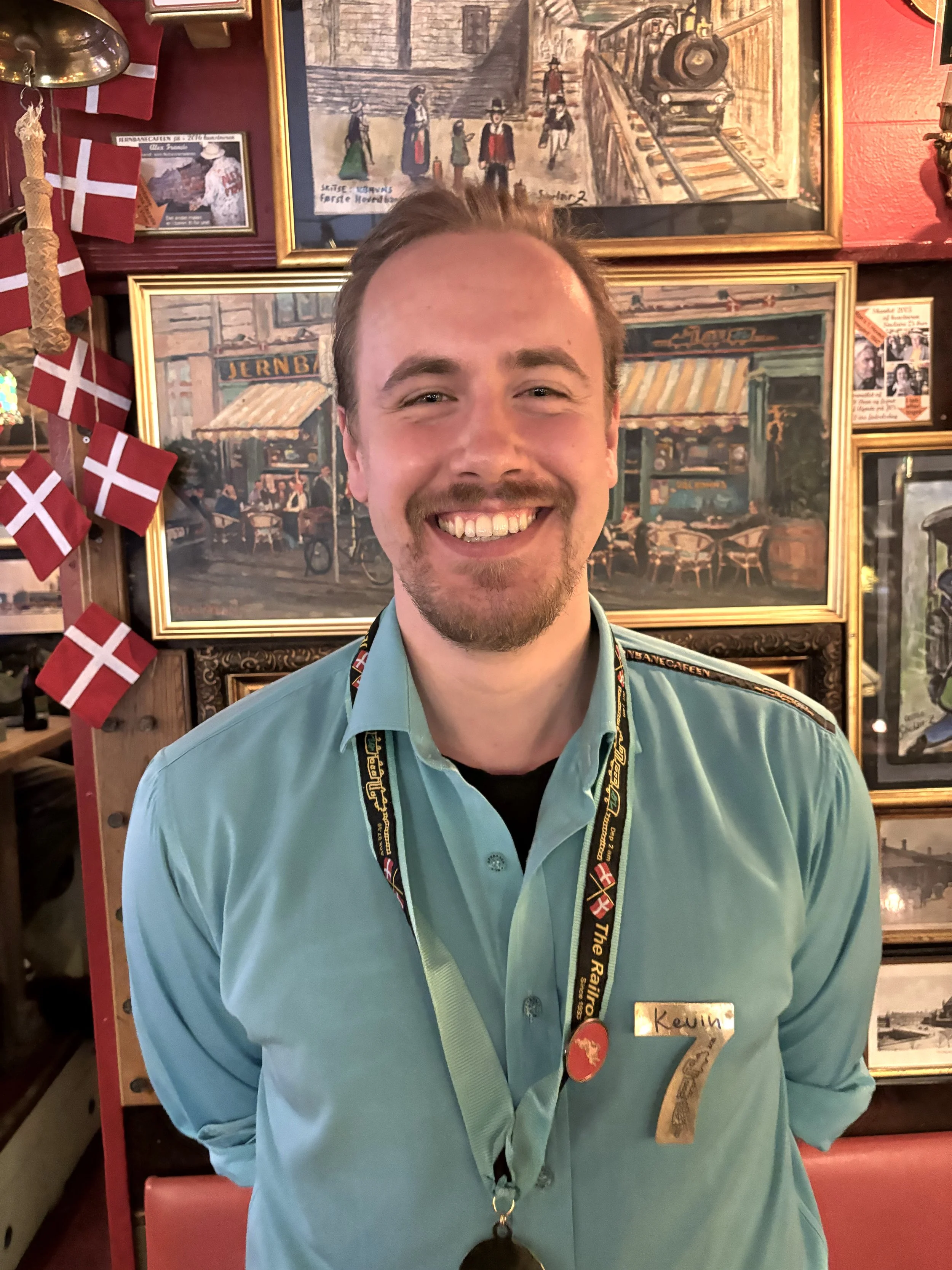 En smilende mand med brun hår og skæg, iført en lyseblå uniform med navnetag, der står "Kevin". Bag ham er en væg fyldt med billeder og billeder af danske motiver og små dannebrogsflag.