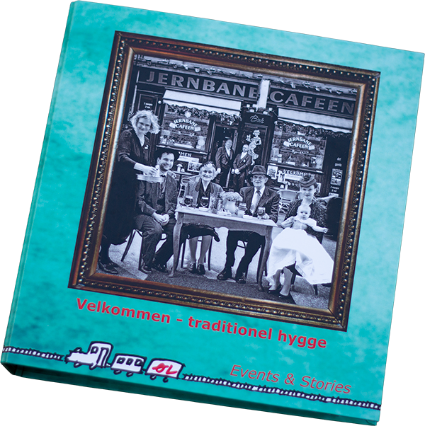 En bog om events og historier med et foto af en familie foran en café, Jernbanecafeen, i sort-hvid. Bogen har en blå cover med rød og sort tekst samt illustrationer af et tog.