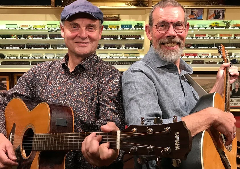 To mænd spiller guitar i en musikbutik med modeltog i baggrunden.