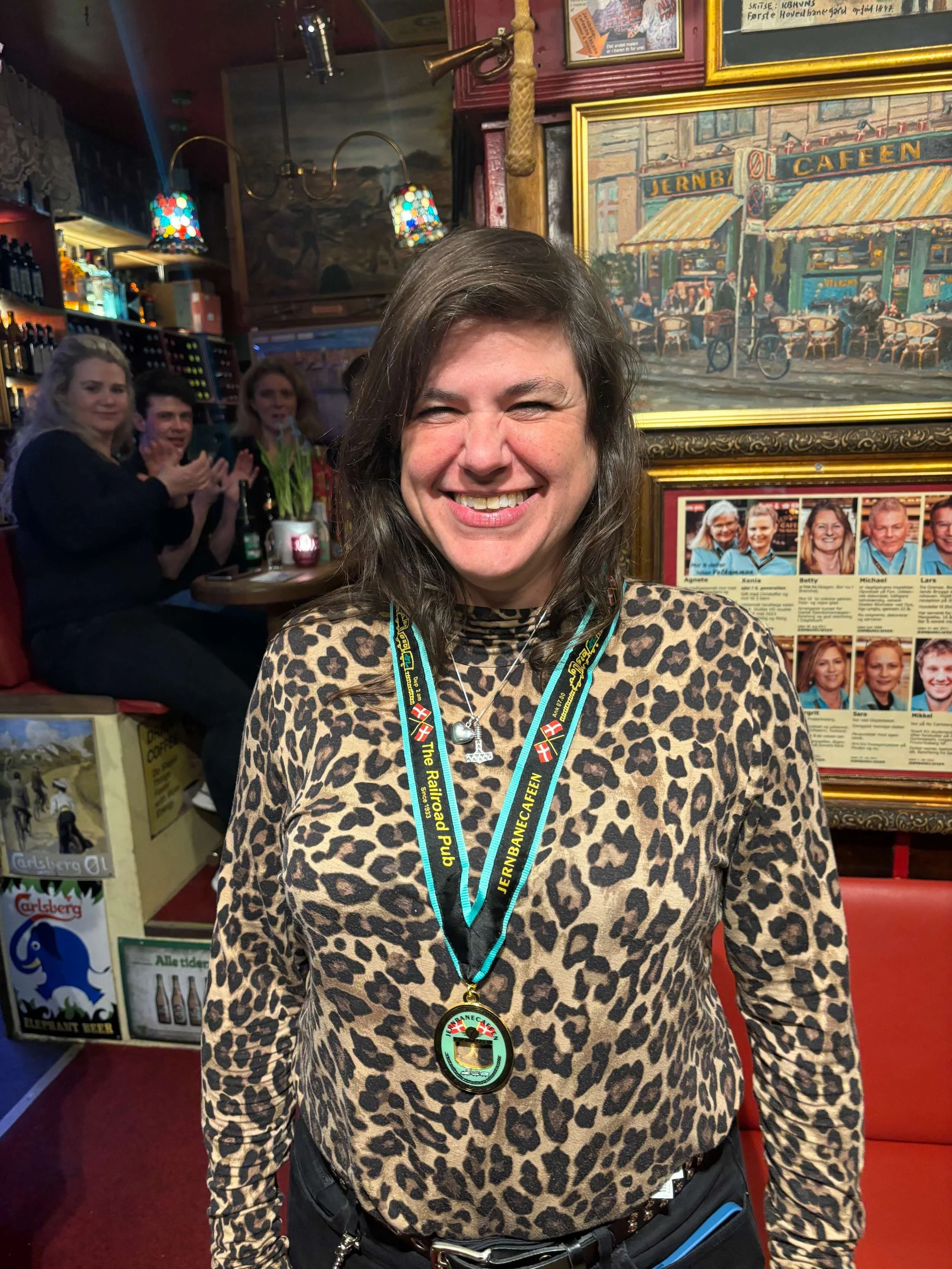 Kvinde i leopardprint bluse med copenhagenløbmedalje rundt halsen, inde i en bar med andre mennesker i baggrunden.