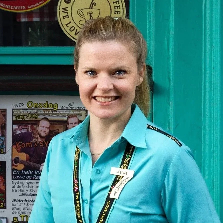 En kvinde med hår sat op i en hestehale, iført lyseblå uniform og navn tag med teksten 'Xenia,' står foran en grøn dør og et avisudklip.
