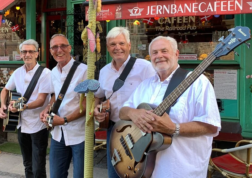 Fire mænd står udenfor Jernbanecaféen, hver med en guitar, smilende.