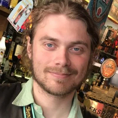 Et close-up portræt af en mand med lyst hår, skæg og blå øjne, på en bar.