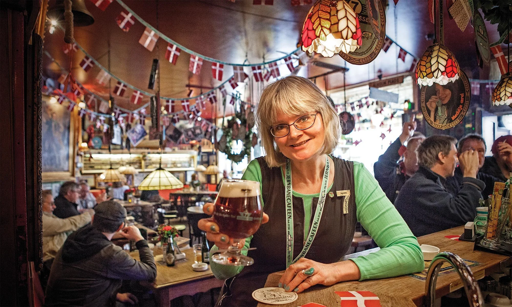 En glad kvinde med briller, siddende ved en bar med en stor glas øl, i en hyggelig bar med dansk flagdekoration.