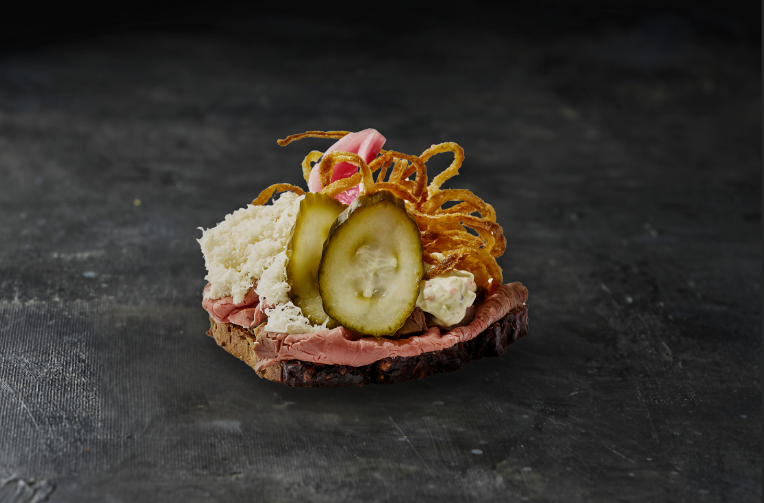 En stykke rugbrød med pålæg, agurk, løg, kød og måske mayonnaise på en mørk baggrund.
