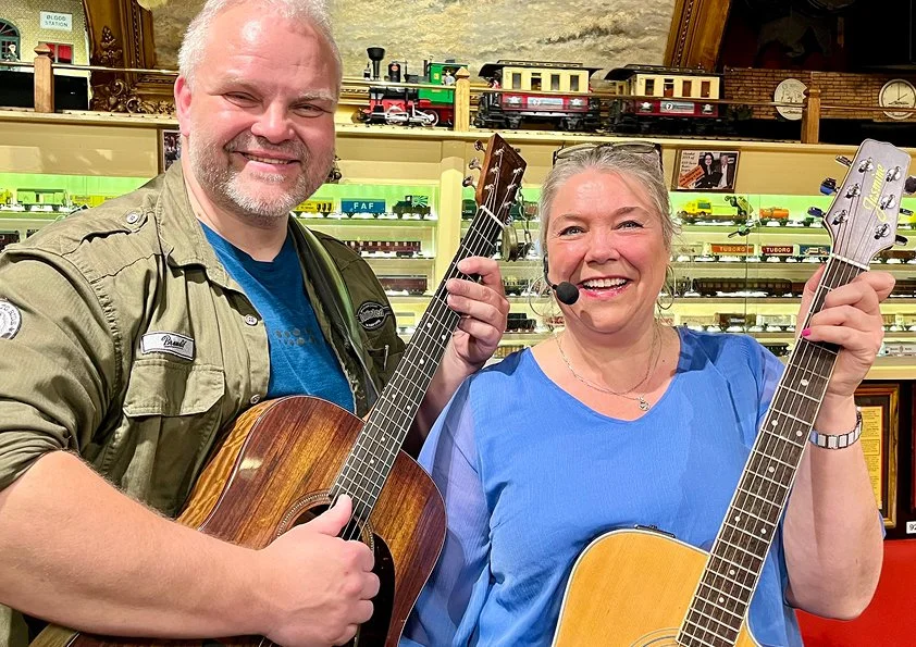 To personer, en mand og en kvinde, smiler og spiller guitar i en modeljernbanebutik med hylder fyldt med modeltog i baggrunden.