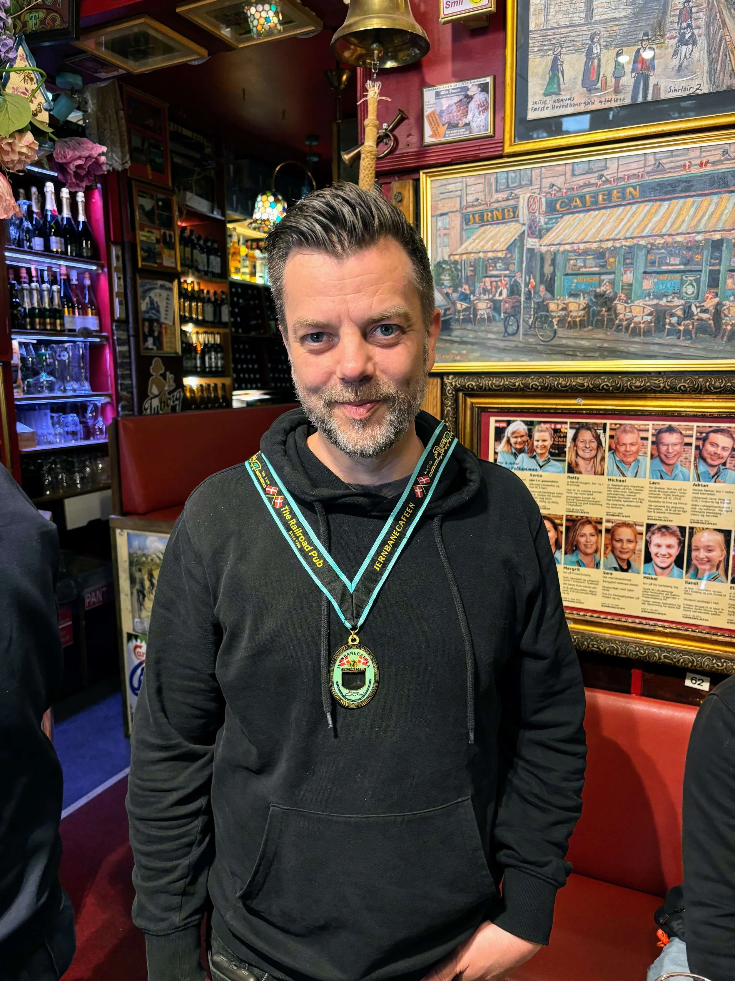 En mand med gråbrunt hår og skæg, iført sort hoodie og et medejerskabsmedalje omkring halsen, står inde i en hyggelig bar eller café med billeder og plakater på væggen.
