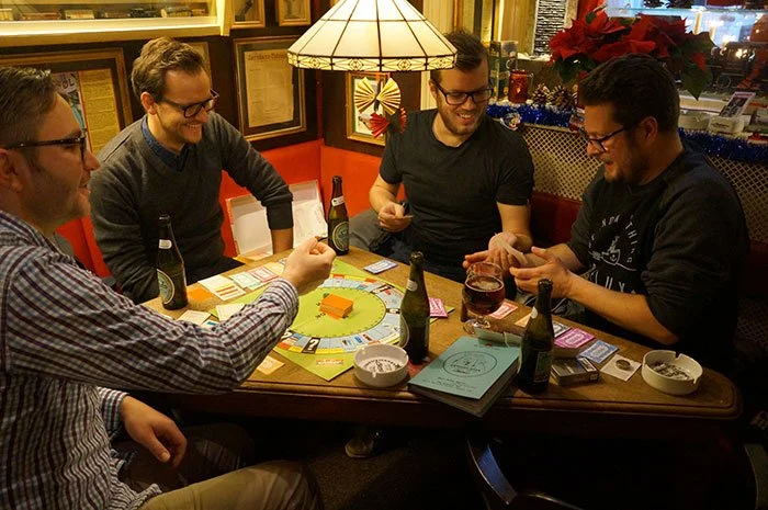 Fem personer spiller et brætspil ved et bord i en indendørs restaurant eller bar, omgivet af julepynt.