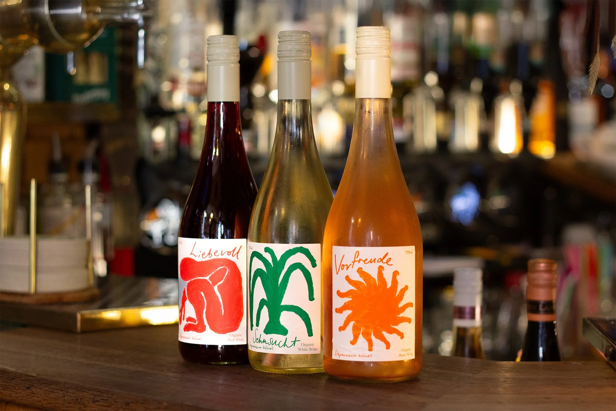 Tre vinflasker med kunstneriske etiketter står på en bar. Baggrunden er sløret med barudstyr og flasker.