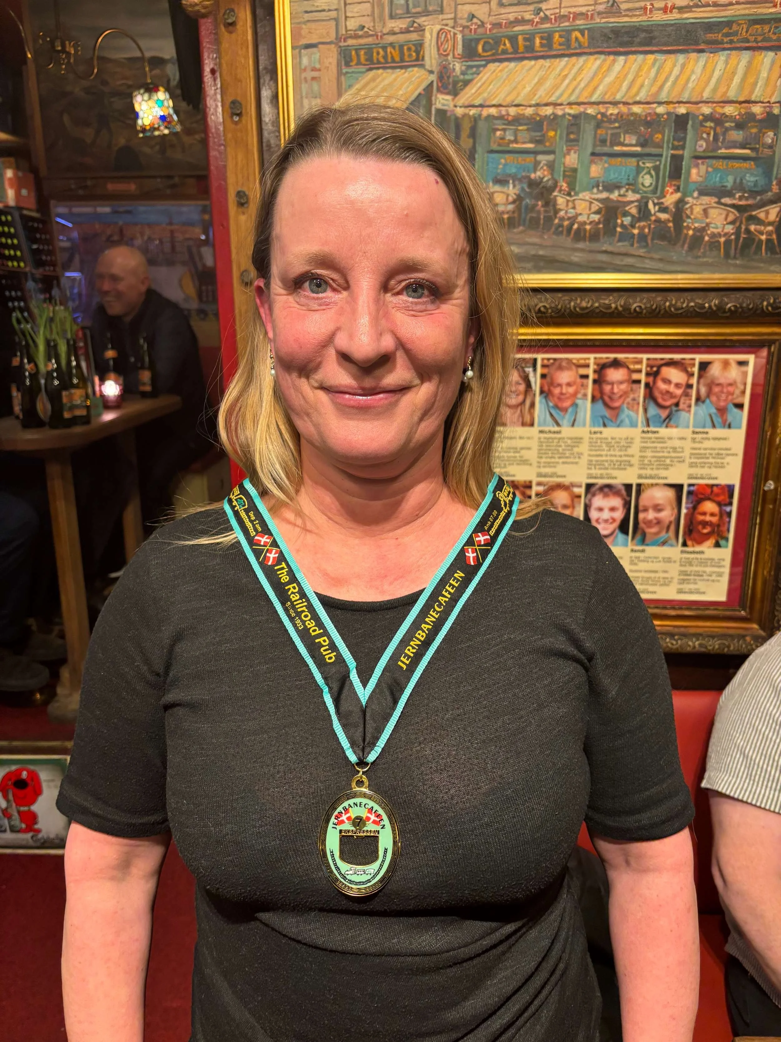 En smilende kvinde iført en medalje og en halskæde med en Jernbanecaféens logo, står i en restaurant eller pub med billeder og tavler på væggene.
