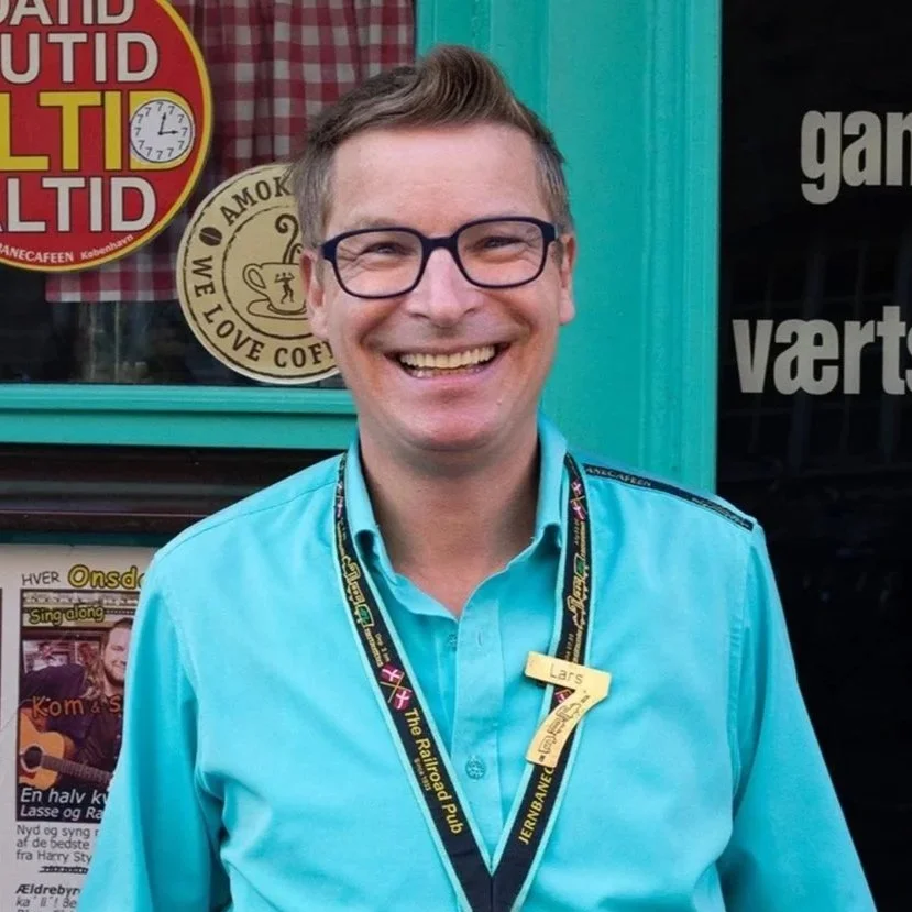 Smilende mand med briller iført en blå skjorte og en lanyard foran en grøn dør, med reklamer og skilt i baggrunden.