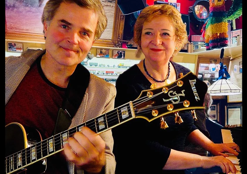To personer sidder ved et klaver. Den ene spiller guitar, og den anden sidder ved klaveret. De er i en hyggelig indendørs setting, måske en café eller musiksted, med farverige dekorationer og billeder på væggene.
