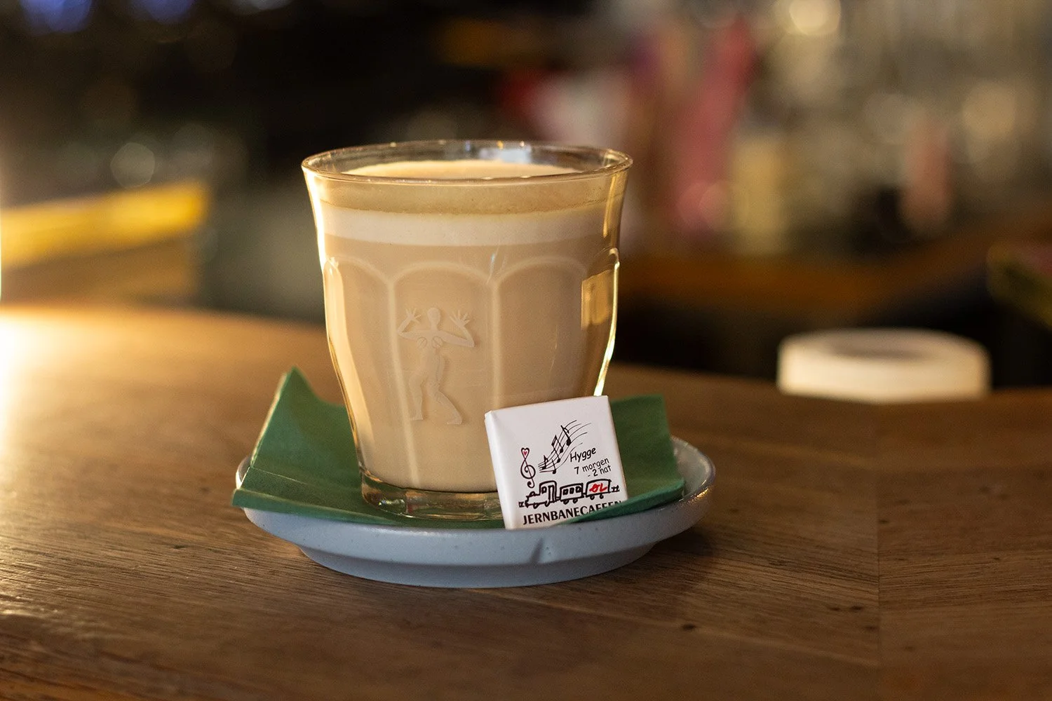 Et glas cappuccino med mælkeskum på en kaffebord, med en lille skål under glasset og en ristret pakke sukker ved siden af, i en café.